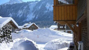 Een houten chalet met een met sneeuw bedekte dak, een houten terras en uitzicht op een besneeerd berglandschap.