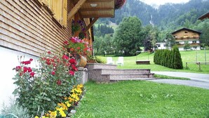 Ein Holzbalkonbau mit einem Blumenbeet voller roter und gelber Blüten, mit Blick auf eine grüne Landschaft, Bäume und einen Berg im Hintergrund.
