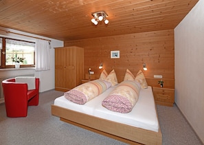 A wooden-paneled room with a bed, a red chair, a window with curtains, and a ceiling light fixture.