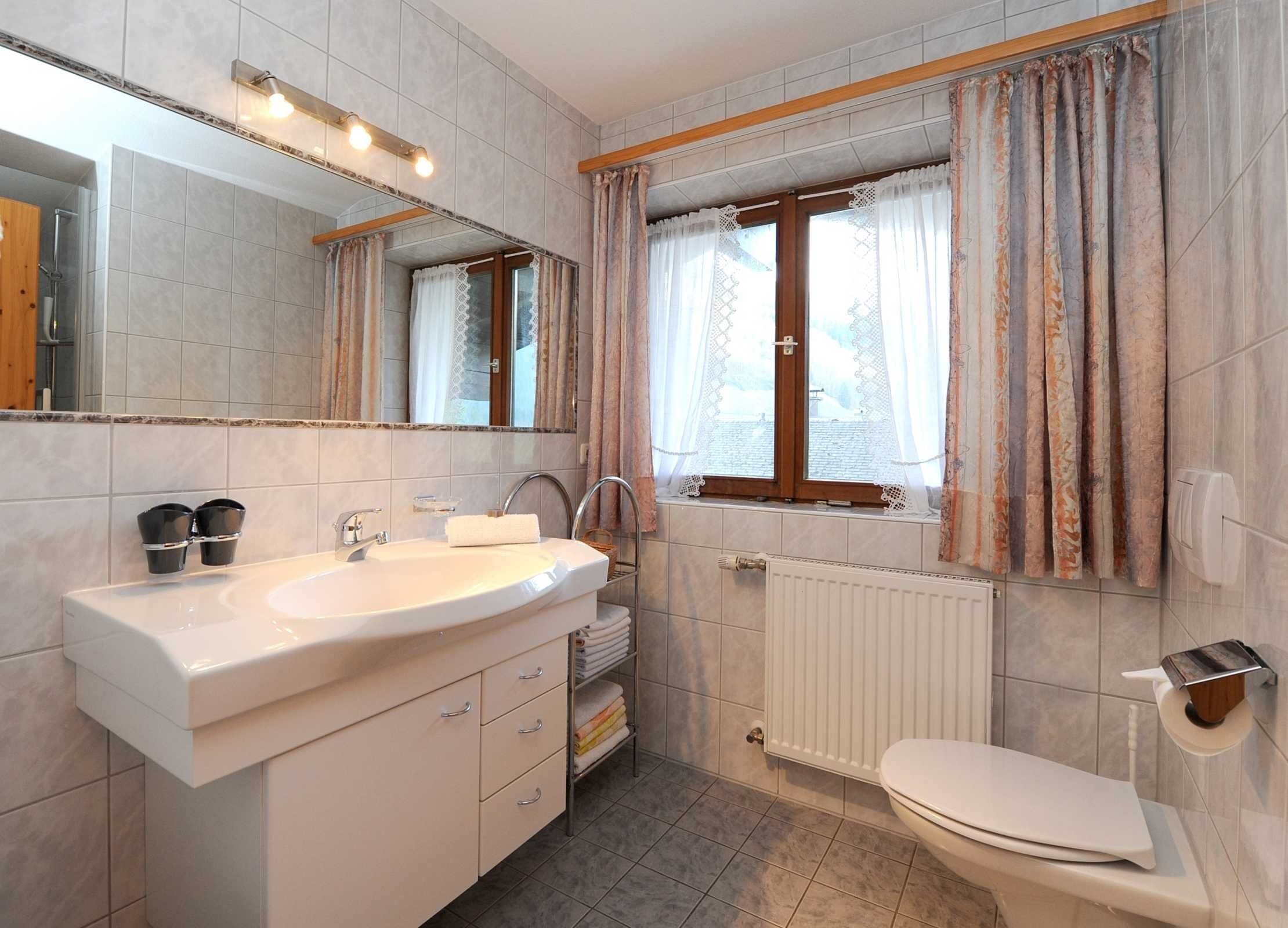 Un baño con lavamanos, espejo, ventana con cortinas, radiador y retrete.