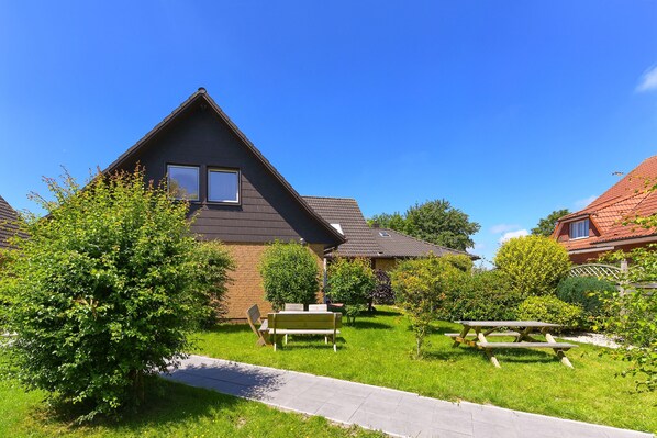 Une maison d’habitation avec un toit en pente, une cour avant avec une table de pique-nique et des bancs, et un ciel bleu dégagé.