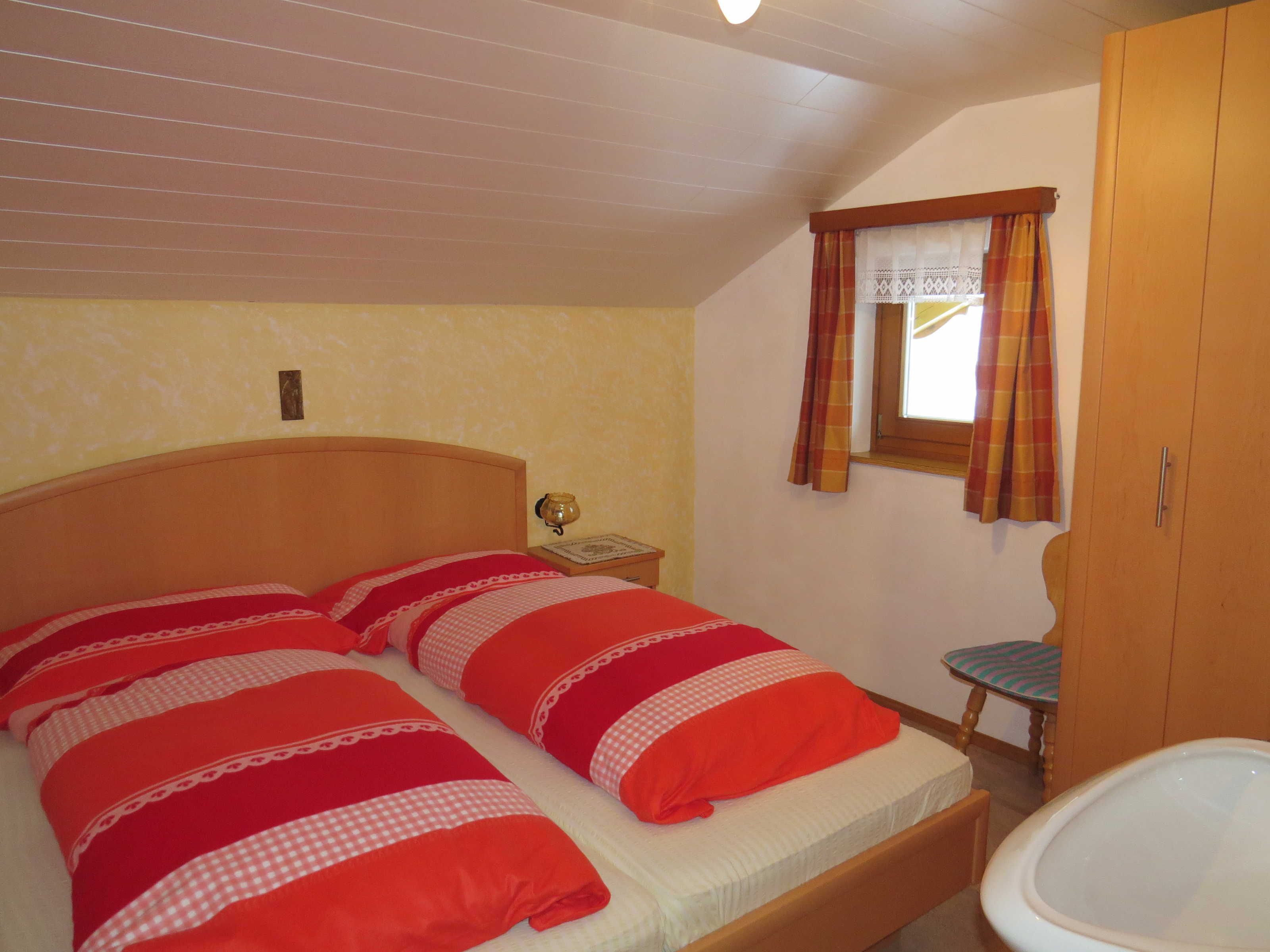 A bedroom with a bed, a nightstand, a window with curtains, and a wooden wardrobe.