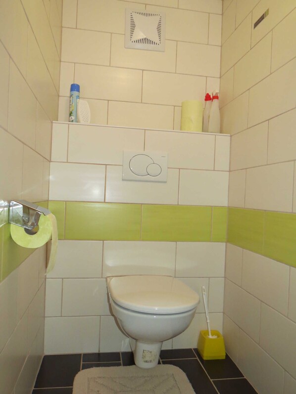 Un baño con inodoro, bidé, dispensador de papel higiénico y estante con artículos de aseo.