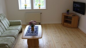 A living room with a green sofa, a wooden coffee table with a vase of flowers, a wooden TV stand with a TV, and two windows with flower boxes. - Cozy accommodation in the Vierseithof in an idyllic, rural location (Crostwitz)