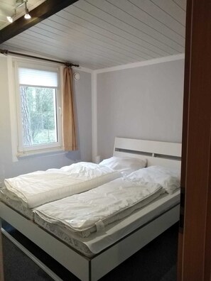 A single-bed room with a window, white walls, and a wooden ceiling.