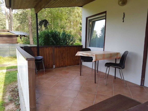 A covered patio area with a table and chairs, a wooden bench, and a view of the surrounding greenery.