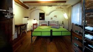 Un dormitorio con una cama verde, suelo de madera, un escritorio con silla, una lámpara y una estantería para vinos.