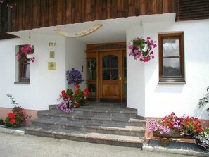 A building with a wooden door, steps, and hanging flower baskets. - Apartment Üntschenspitze - Apartments Haus Monika (Rehmen)