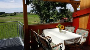 Un balcon avec une table et des chaises, donnant sur un champ verdoyant parsemé d’arbres et sous un ciel dégagé.
