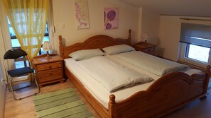 A double bed with wooden headboard, two bedside tables, a nightstand, a chair, a green striped rug, and a window with blinds. - Apartment Gold - Ferienhof Gold (Waischenfeld)