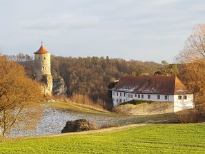 A castle with a tower, a white building with a red roof, and a river in the foreground. - Apartment Gold - Ferienhof Gold (Waischenfeld)