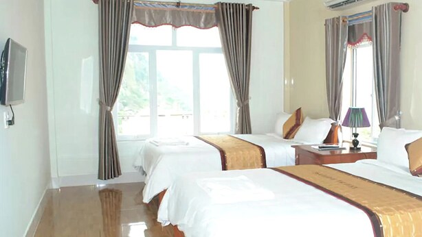 A hotel room with two beds, a flat-screen TV, a bedside table with a lamp, and a window with curtains.
