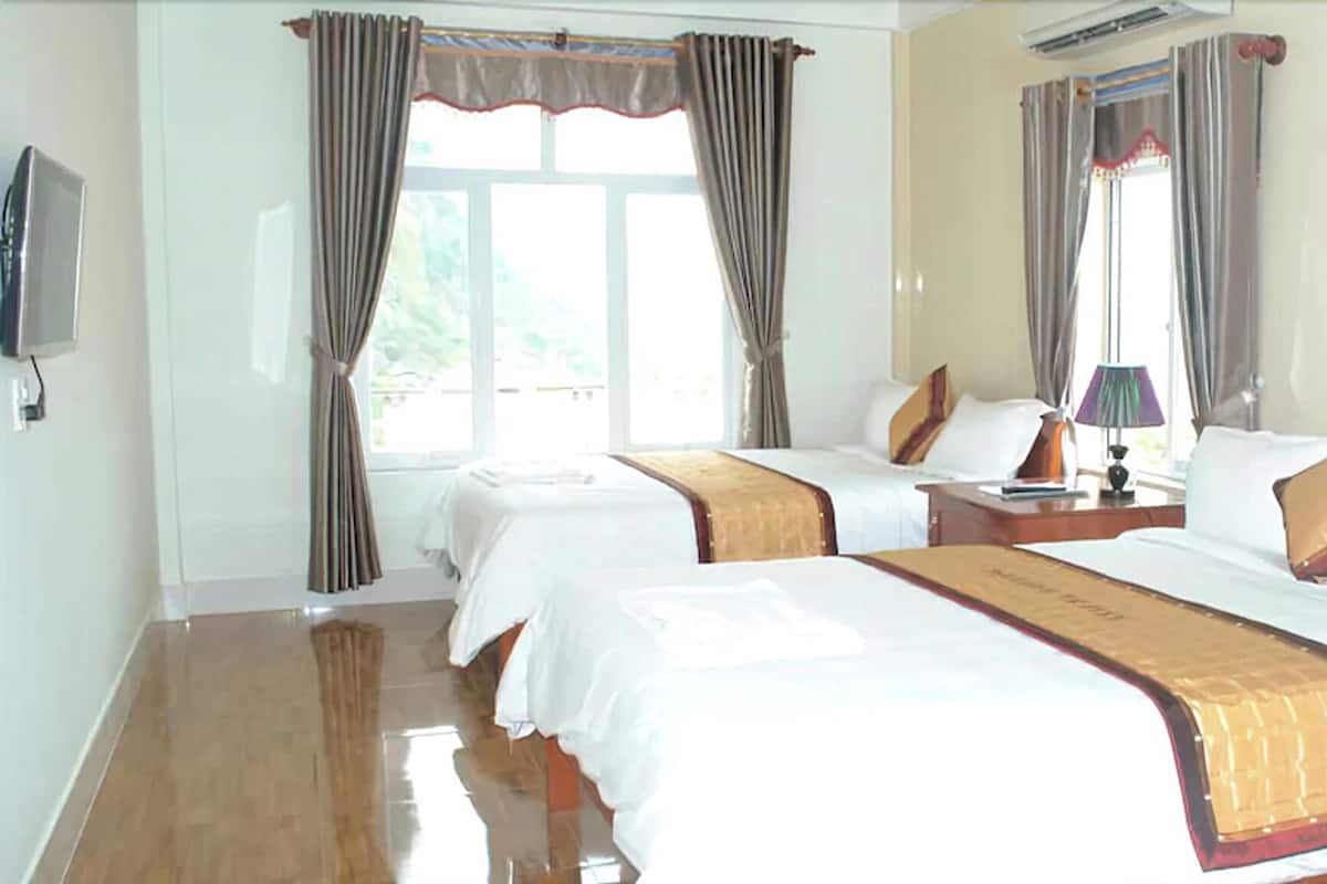 A hotel room with two beds, a flat-screen TV, a bedside table with a lamp, and a window with curtains.