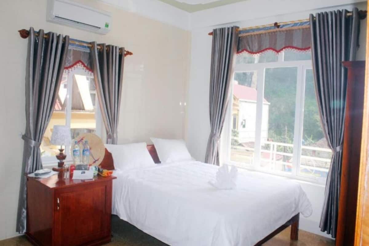 A hotel room with a bed, a wooden nightstand with a lamp, and curtains.