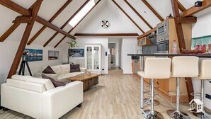 Amplia sala de estar con techo alto, vigas de madera, un sofá blanco, una mesa de centro y una zona de cocina con taburetes de bar.