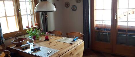 A wooden dining table with chairs, a lamp, a vase with flowers, and a window with curtains.