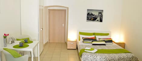 A bedroom with a bed, bedside tables, a desk with chairs, and a door.