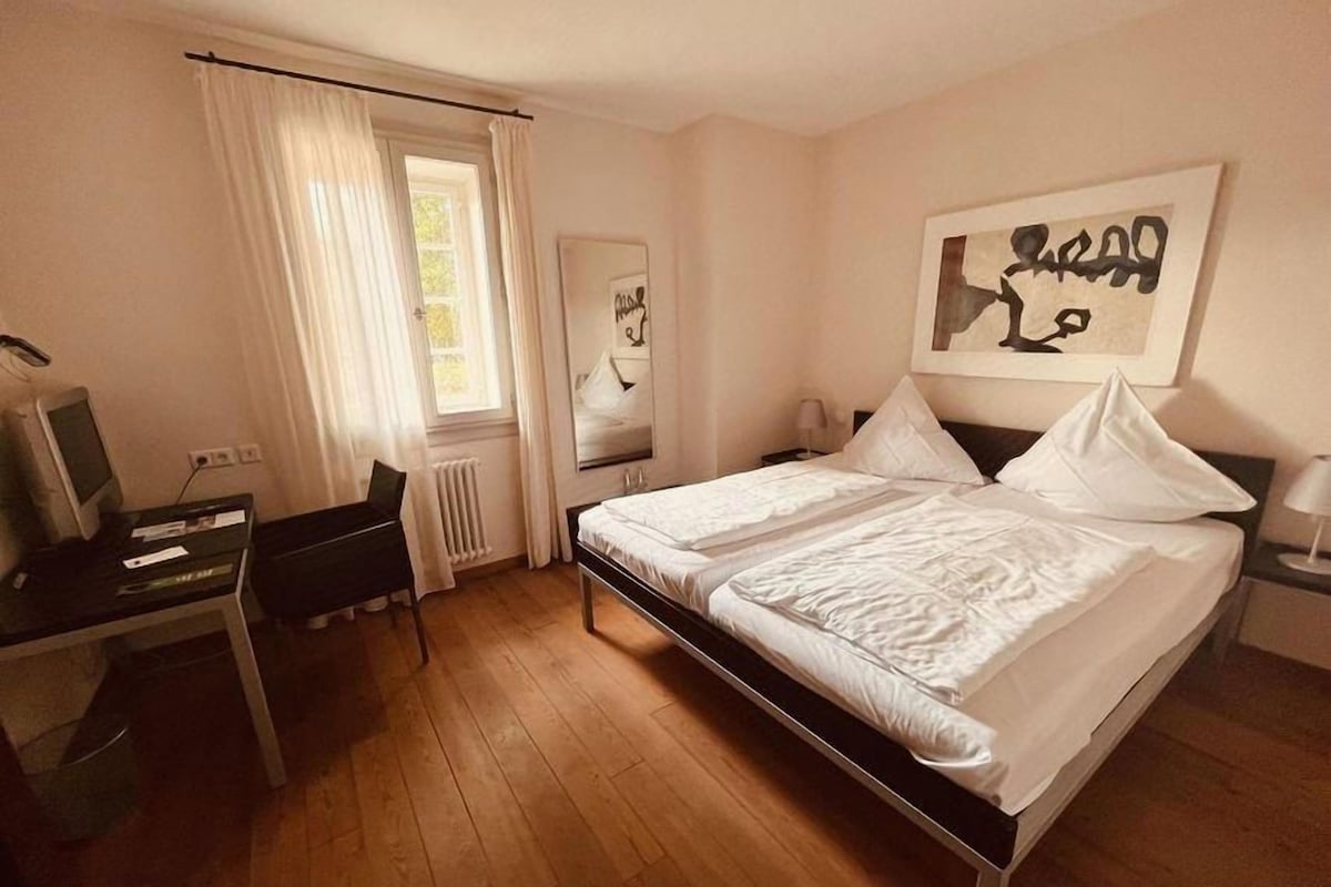 Ein Hotelzimmer mit Bett, Schreibtisch, Stuhl, Heizkörper und einem fenster mit Vorhängen.