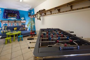 A room with a foosball table, a sofa with a blue and yellow checkered cushion, a small round table with a green stool, and a window with a blue curtain. - Top 1 - 3 (2) - Apartment block Charisma (Fieberbrunn)