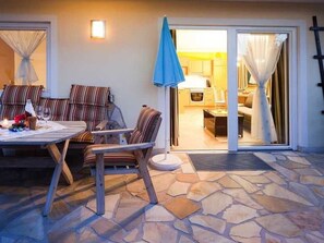 Eine Terrasse mit Steinboden, einem Tisch mit Stühlen und Blick in die Küche durch eine Glastür.