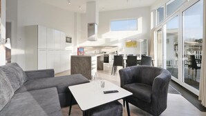 A modern living space with a grey sofa, a white coffee table, and a grey armchair. There is a kitchen area in the background with white cabinets and a dining area with a table and chairs.