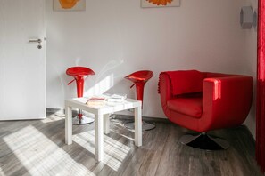 Une pièce avec une porte blanche, une table blanche sur laquelle se trouvent un livre et une boîte, deux chaises rouges et un canapé rouge.