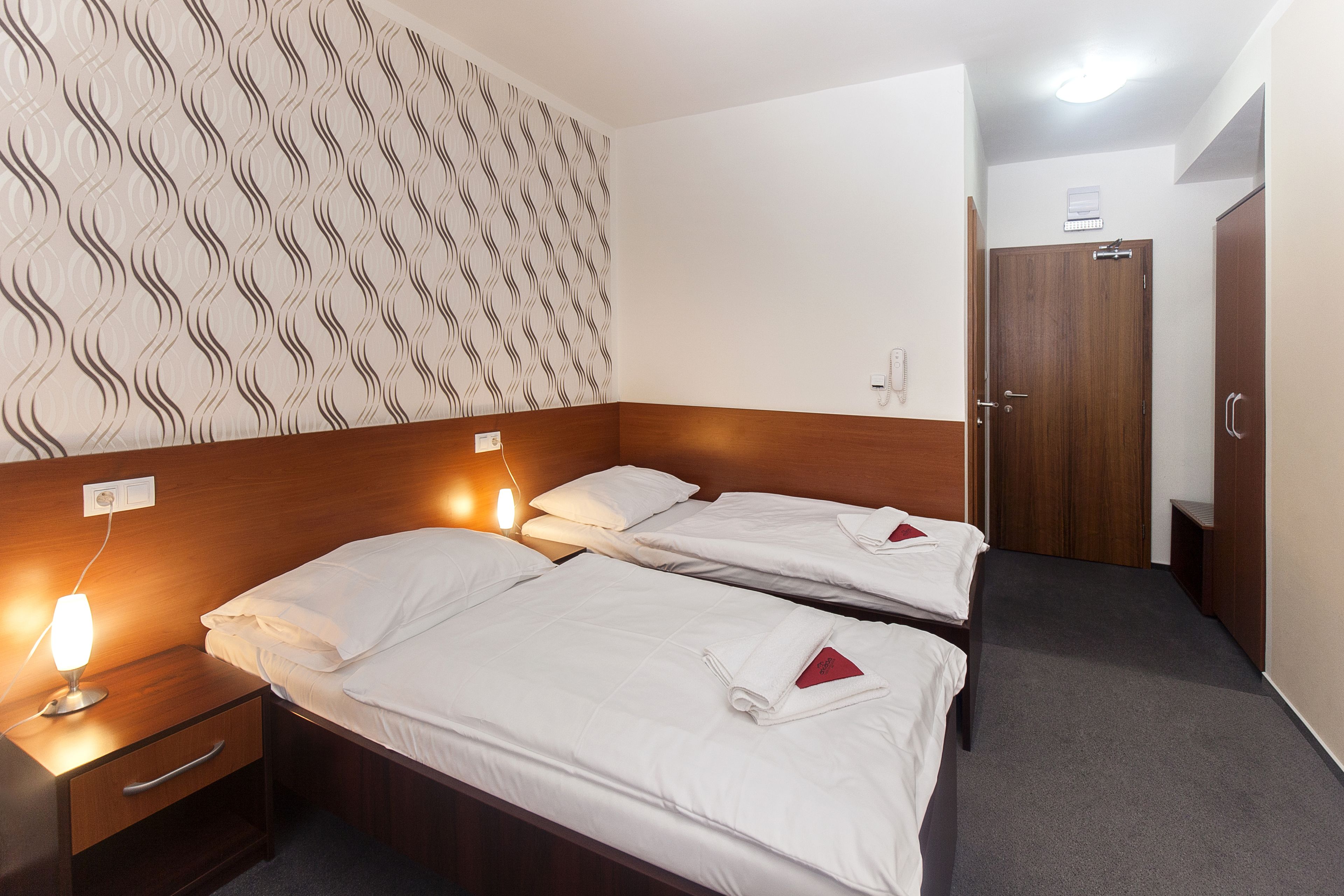 Habitación de hotel con dos camas individuales, cabecero de madera, pared estampada, mesita de noche con lámpara y una puerta a la derecha.