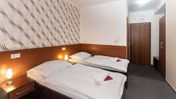 Habitación de hotel con dos camas individuales, cabecero de madera, pared estampada, mesita de noche con lámpara y una puerta a la derecha.