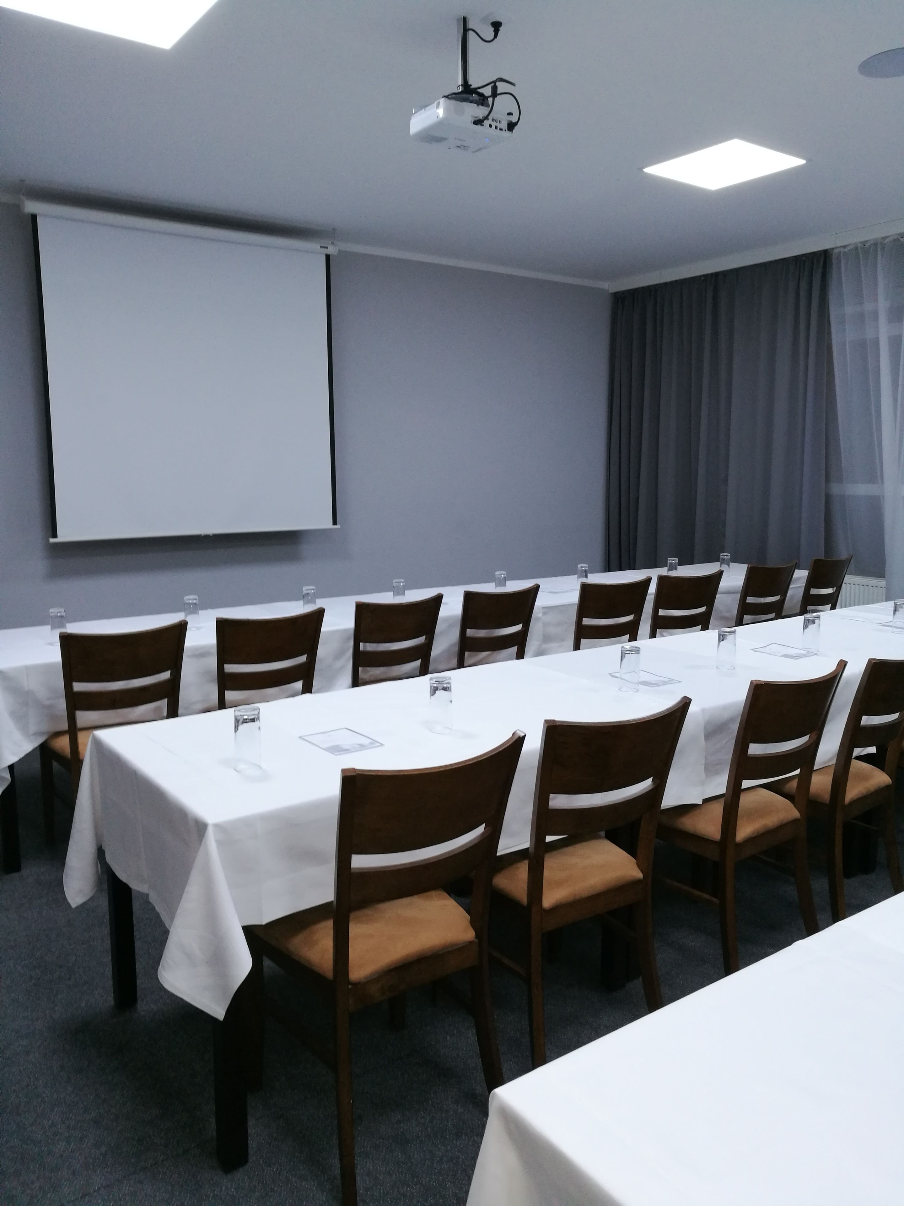 Une salle de conférence équipée de plusieurs tables et chaises, d’un écran de projection et d’une grande fenêtre avec des rideaux.