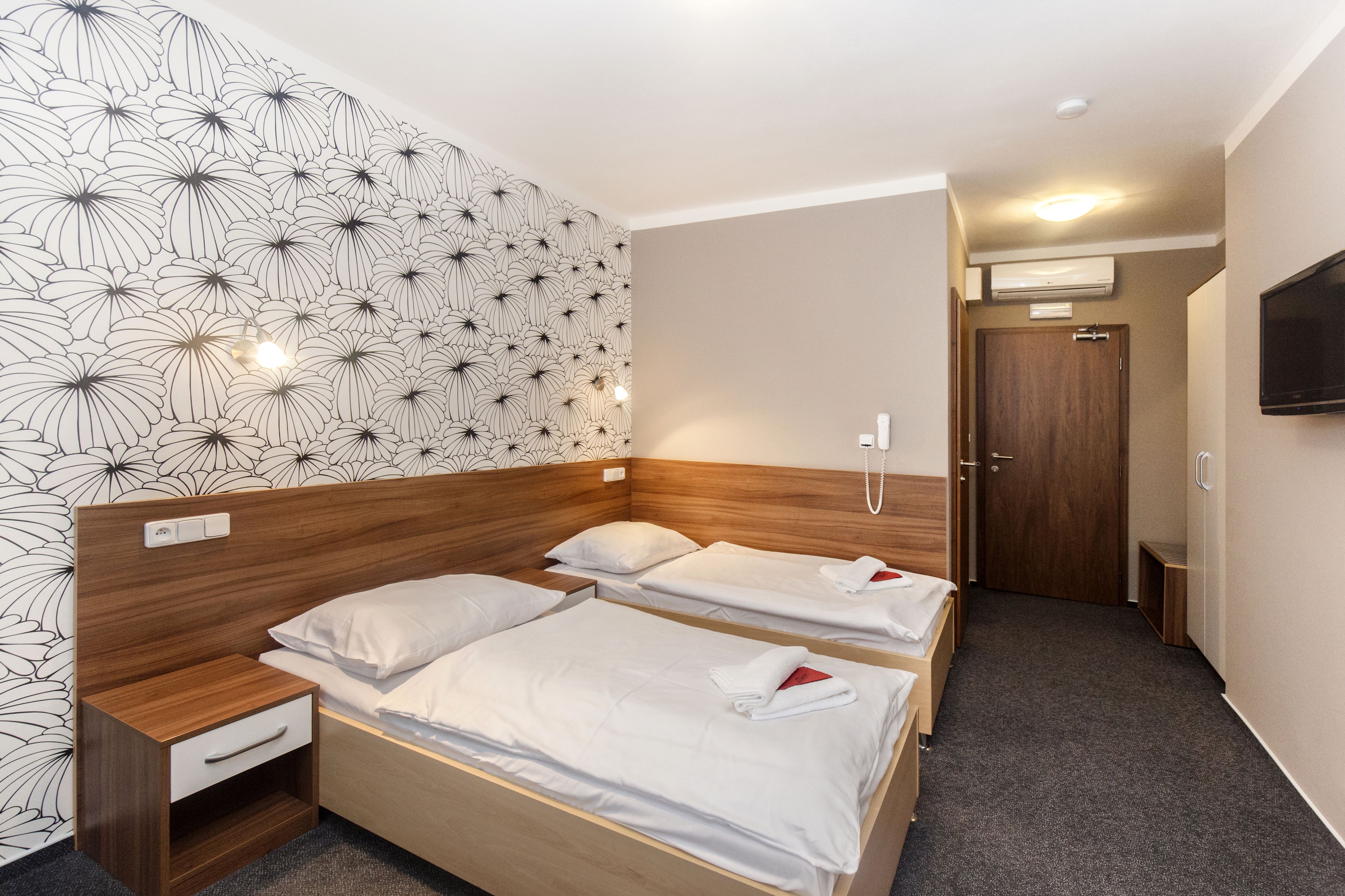 Habitación de hotel con dos camas, cabecera de madera, mesita de noche, televisor montado en la pared y puerta con equipo de aire acondicionado.