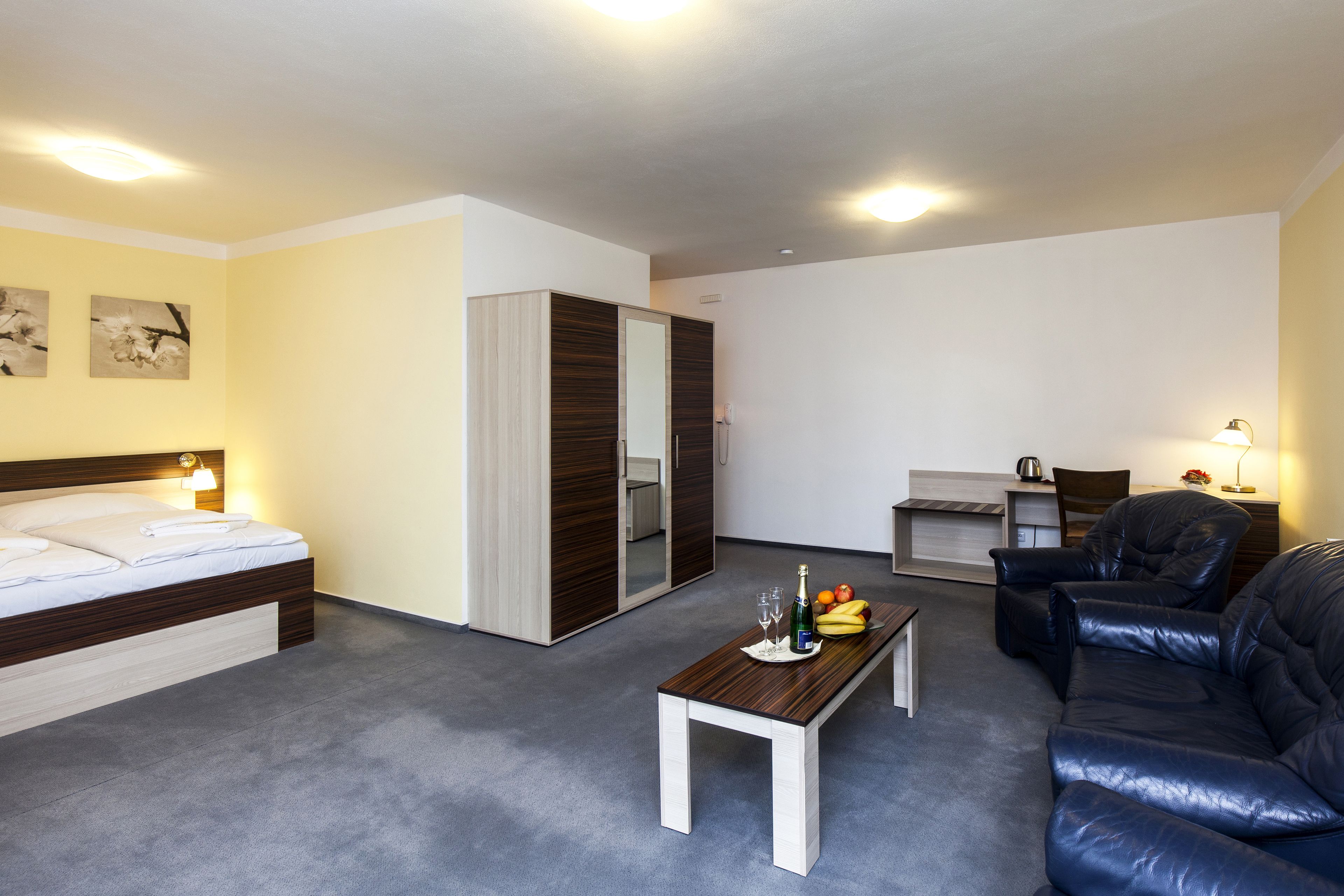 A modern hotel room with a bed, a sofa, a dining table with fruit and drinks, and a desk with a chair.