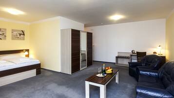 A modern hotel room with a bed, a sofa, a dining table with fruit and drinks, and a desk with a chair.