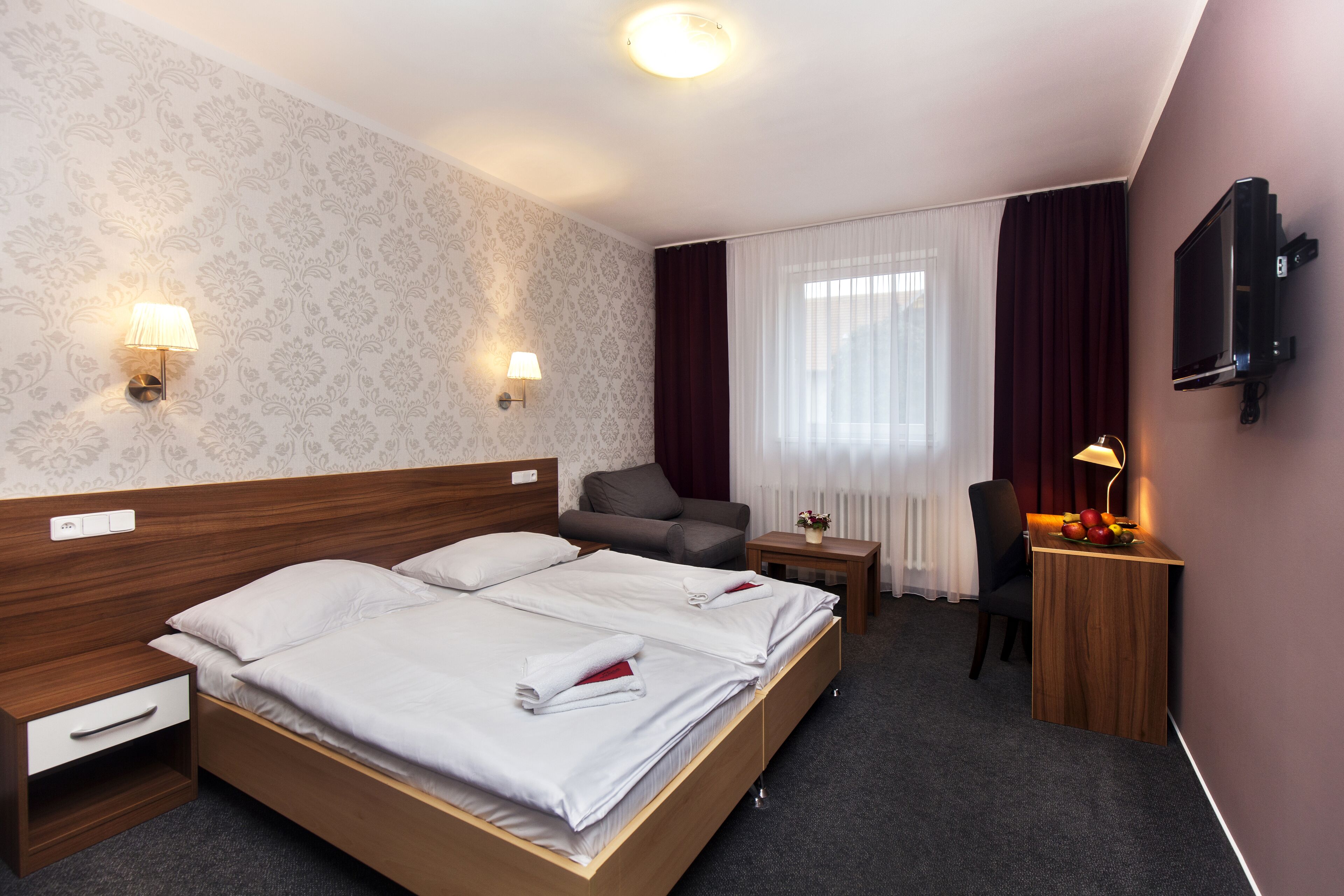 A hotel room with a large bed, two bedside tables, a desk with a chair, a TV mounted on the wall, and a window with curtains.