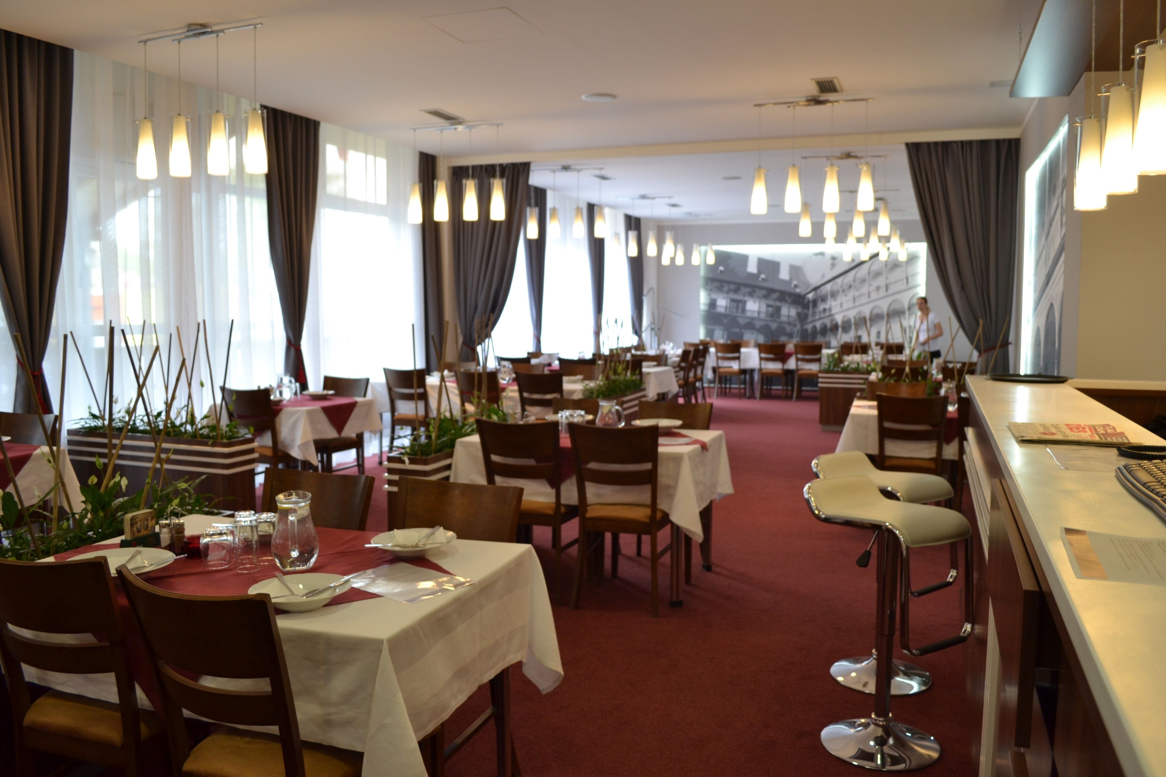Un restaurante con varias mesas y sillas, algunas con manteles blancos y otras con rojos. Hay luces colgantes sobre las mesas, y la sala tiene ventanas amplias con cortinas.