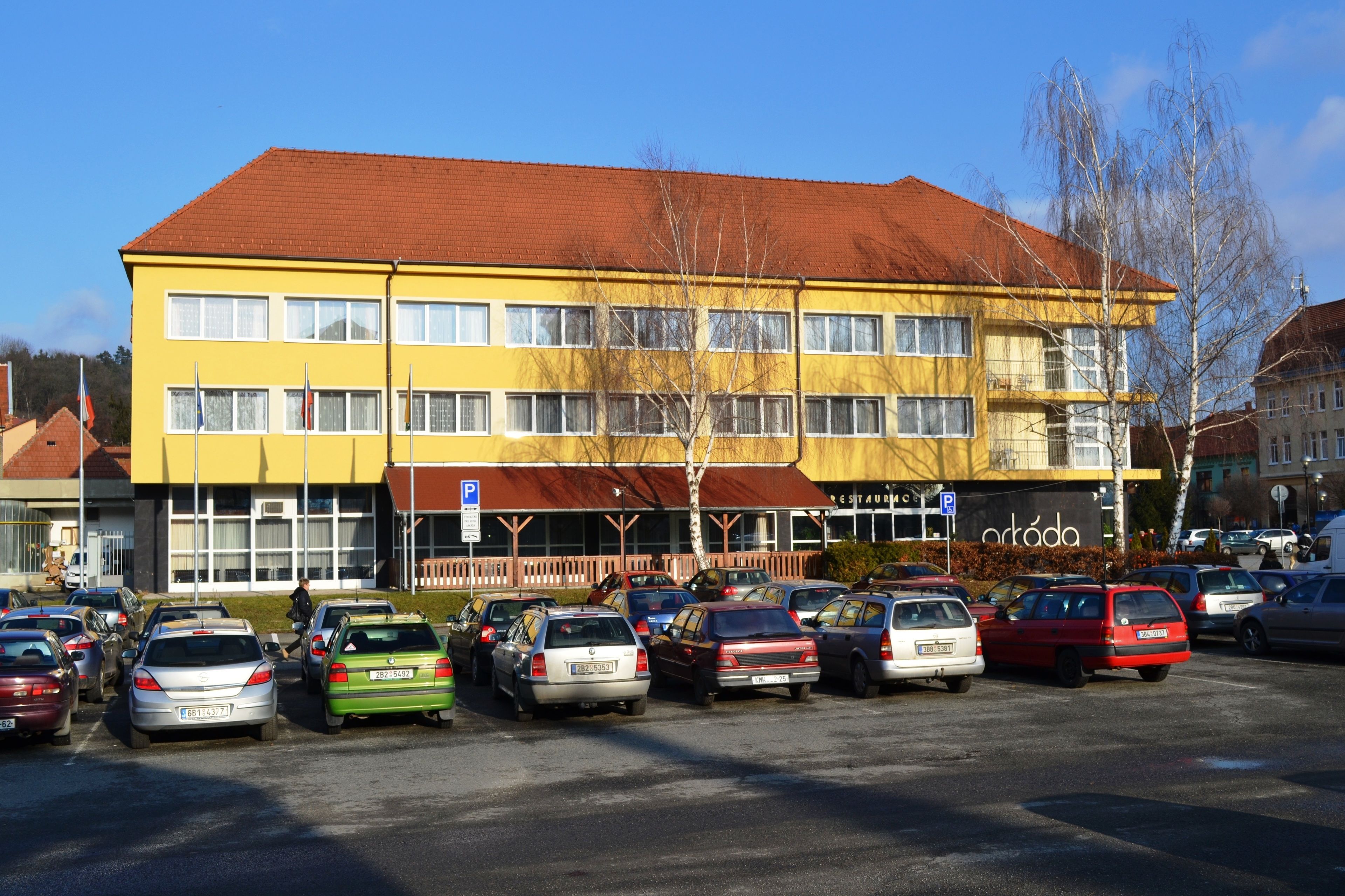 Ein mehrstöckiges, gelbes Gebäude mit rotem Dach, ein Parkplatz mit verschiedenen Autos und ein Schild für ein Restaurant namens "ortada" im Erdgeschoss.