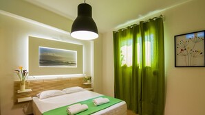 A neatly arranged hotel room with a bed, a bedside table with a lamp, a window with curtains, and a framed picture on the wall. - Castello Di Porto (Mylopotamos)