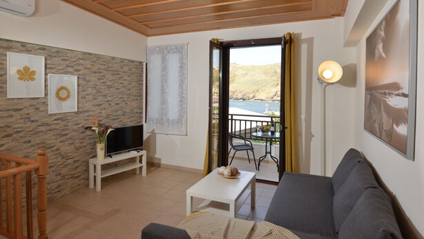 A living room with a stone wall, a television, a sofa, a small table, and a balcony with a view of the sea. - Castello Di Porto (Mylopotamos)
