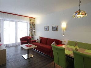 A living room with a red sofa, green chairs, a coffee table, and a dining table. There are two wall-mounted picture frames and a hanging lamp.