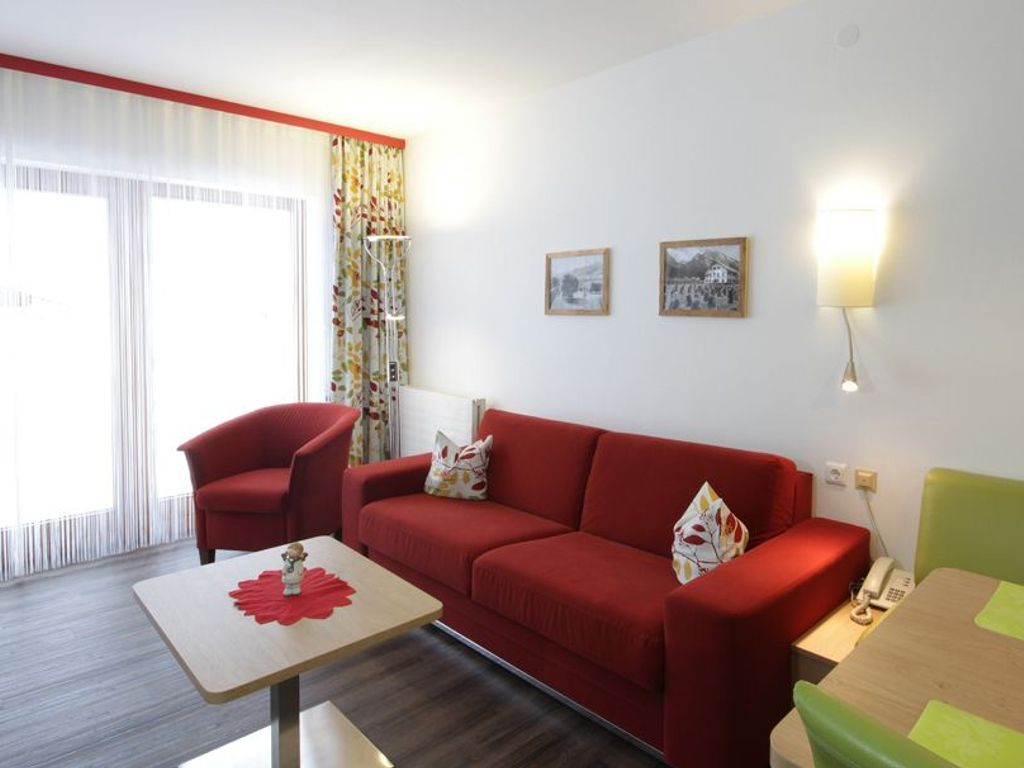 Uma sala de estar moderna com um sofá vermelho, uma mesa pequena e uma cadeira verde. Há duas imagens emolduradas na parede e um abajur na mesa lateral.