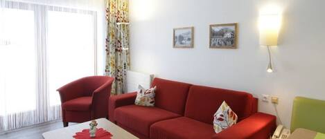 Uma sala de estar moderna com um sofá vermelho, uma mesa pequena e uma cadeira verde. Há duas imagens emolduradas na parede e um abajur na mesa lateral.