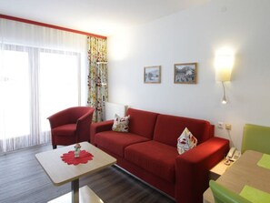 A modern living room with a red sofa, a small table, and a green chair. There are two framed pictures on the wall and a lamp on the side table.