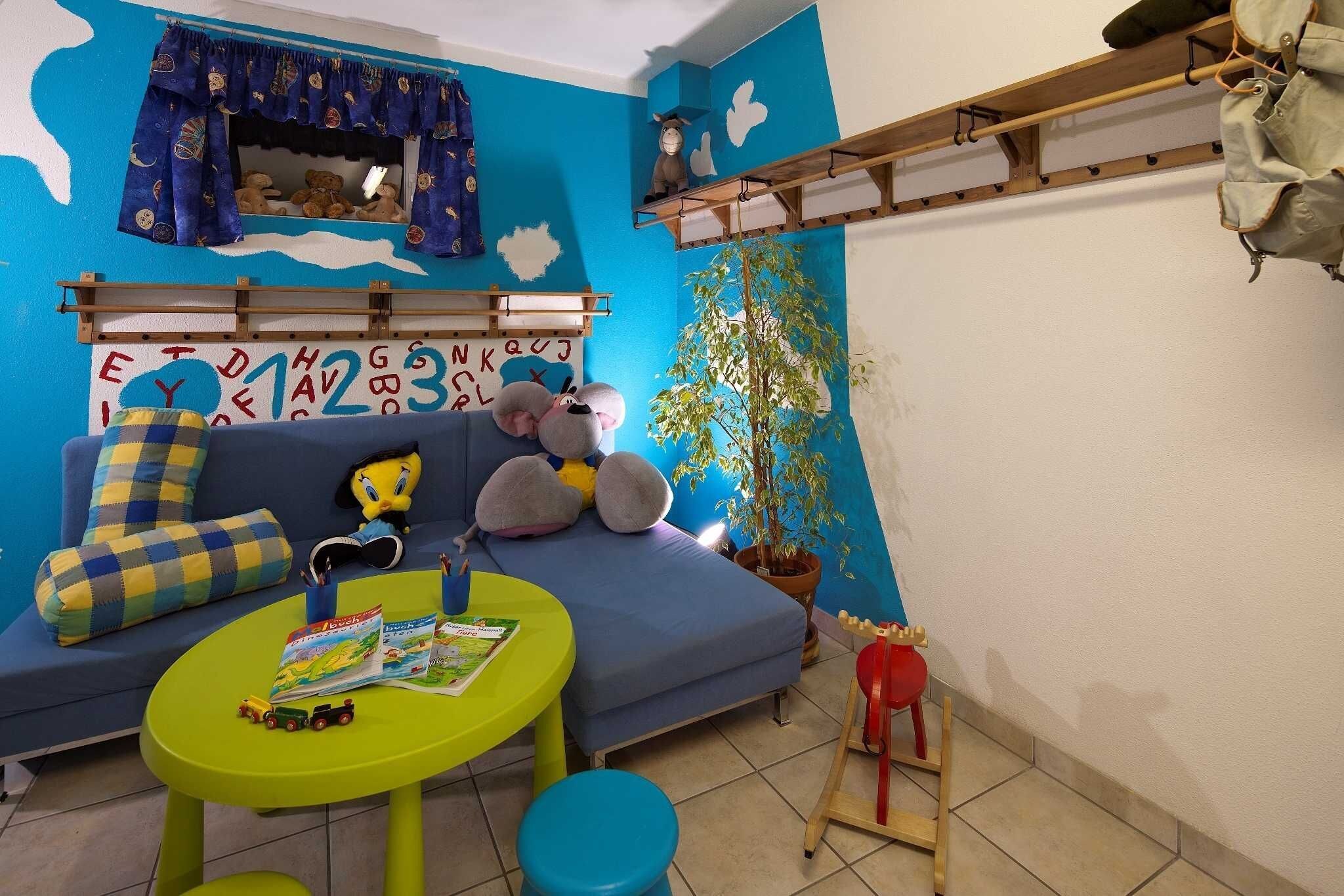 A room with a blue sofa, a green table, a window with curtains, a shelf with items, and a potted plant.