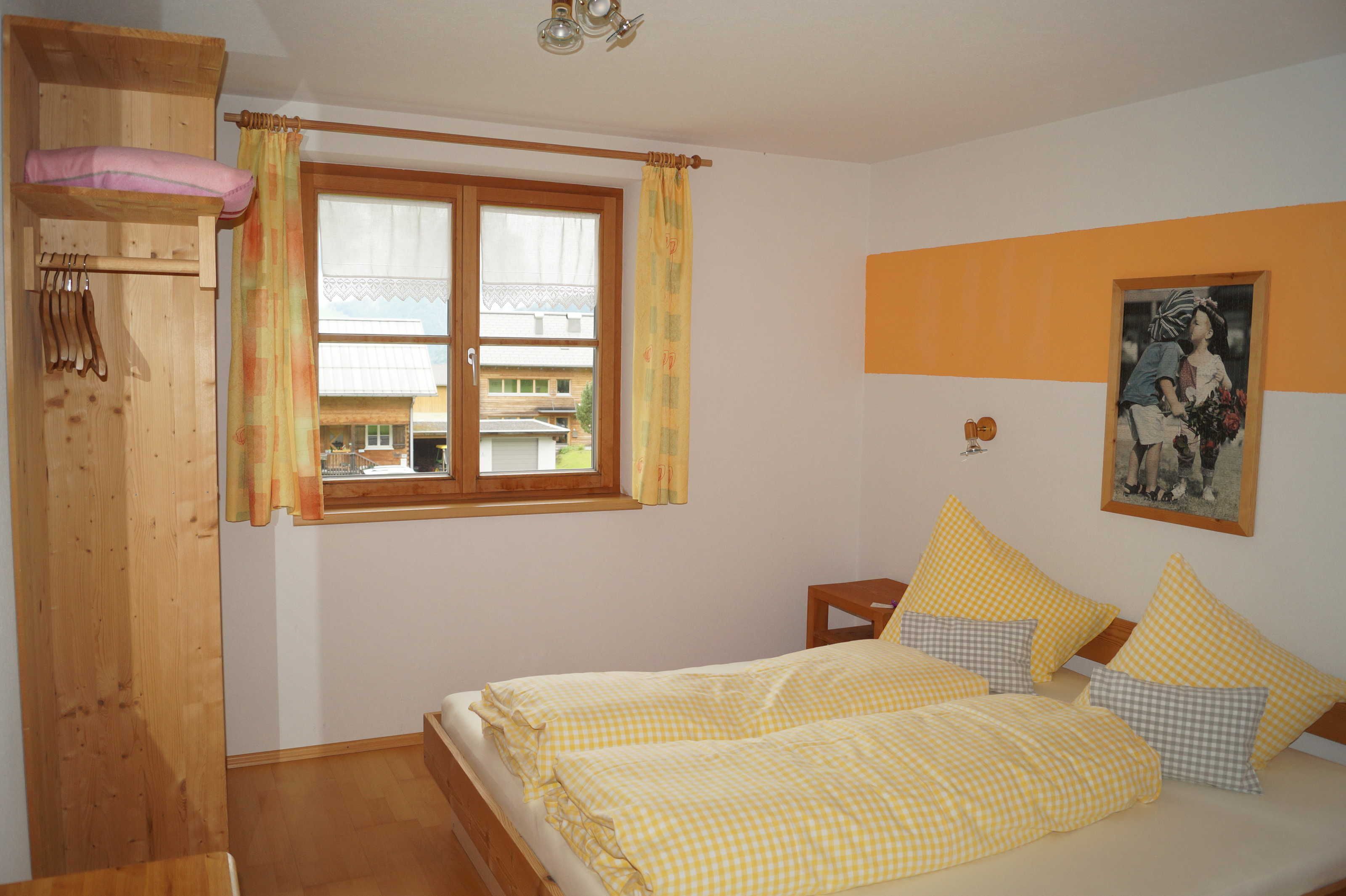 Een slaapkamer met een bed, een houten garderobe, een raam met gordijnen, een nachtkastje en een aan de muur hangend schilderij.