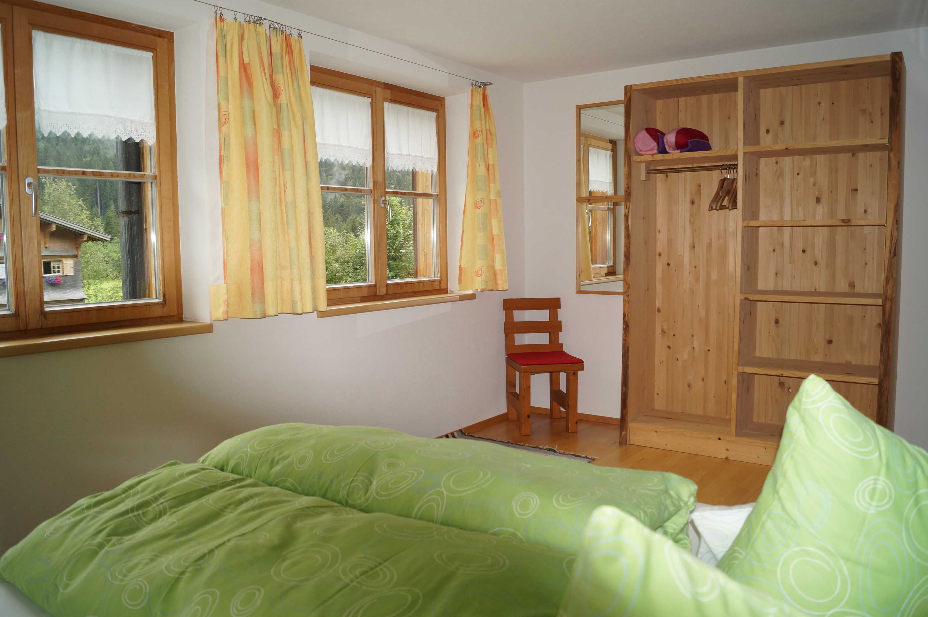 Un dormitorio con cama, una silla, un armario de madera y una ventana con cortinas.