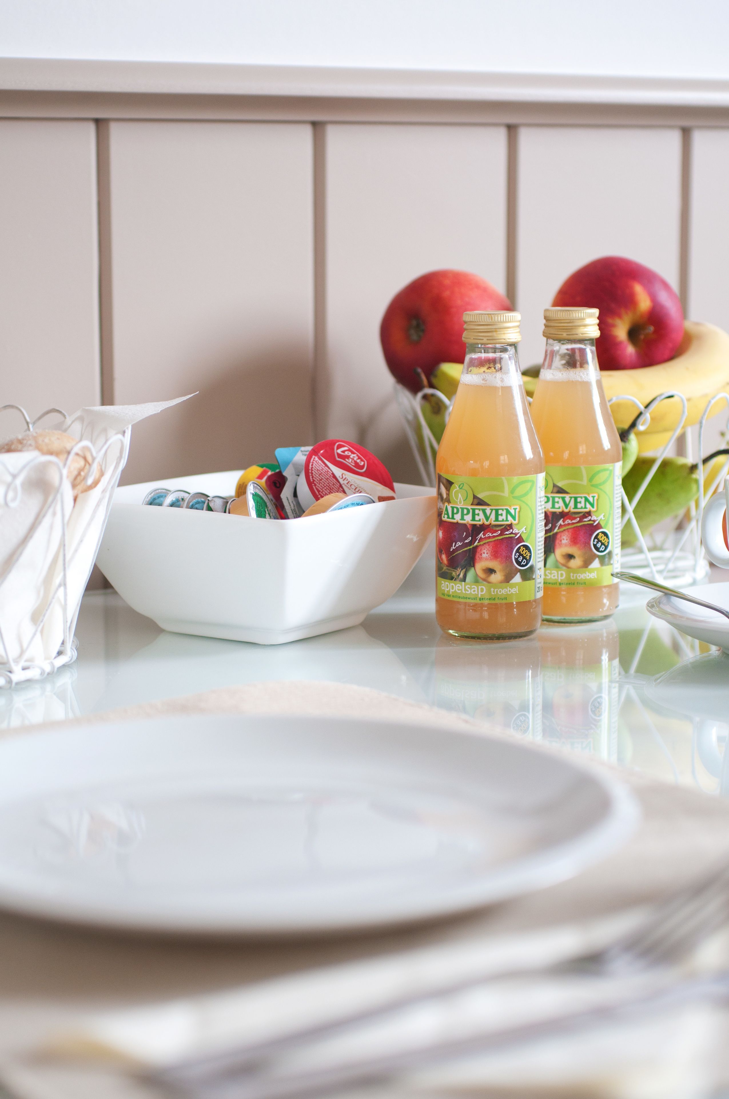 A table with two bottles of apple cider, a bowl of snacks, and a fruit basket with apples.