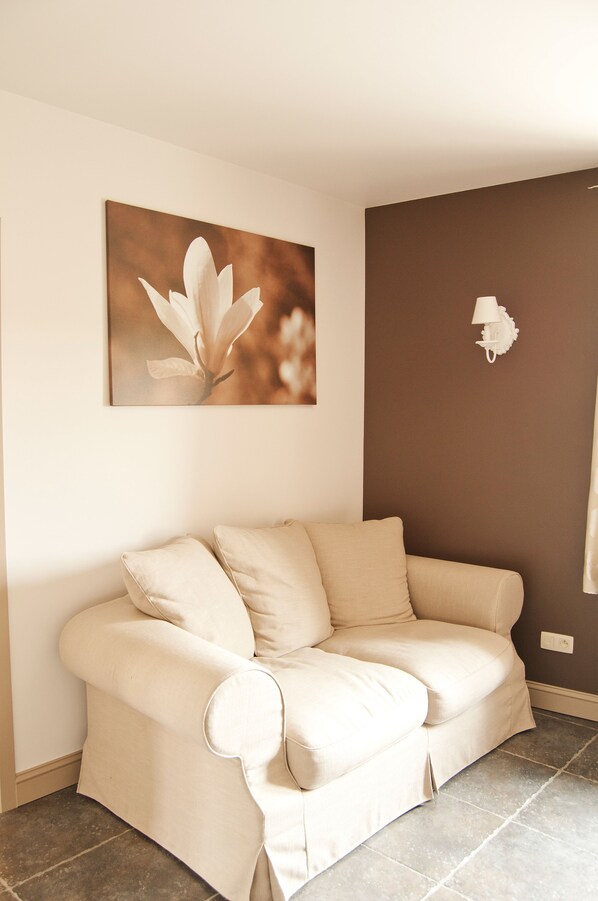 A beige sofa with a cushion, a wall-mounted picture of a flower, and a white wall-mounted lamp. - De Fruithoeve (Hoeselt)