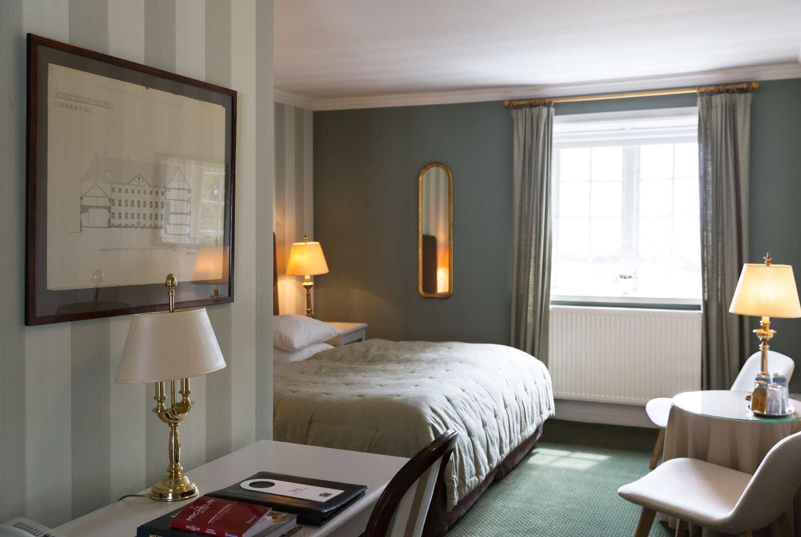 A hotel room with a bed, two bedside tables with lamps, a desk with a chair, a framed architectural drawing, and a window with curtains.