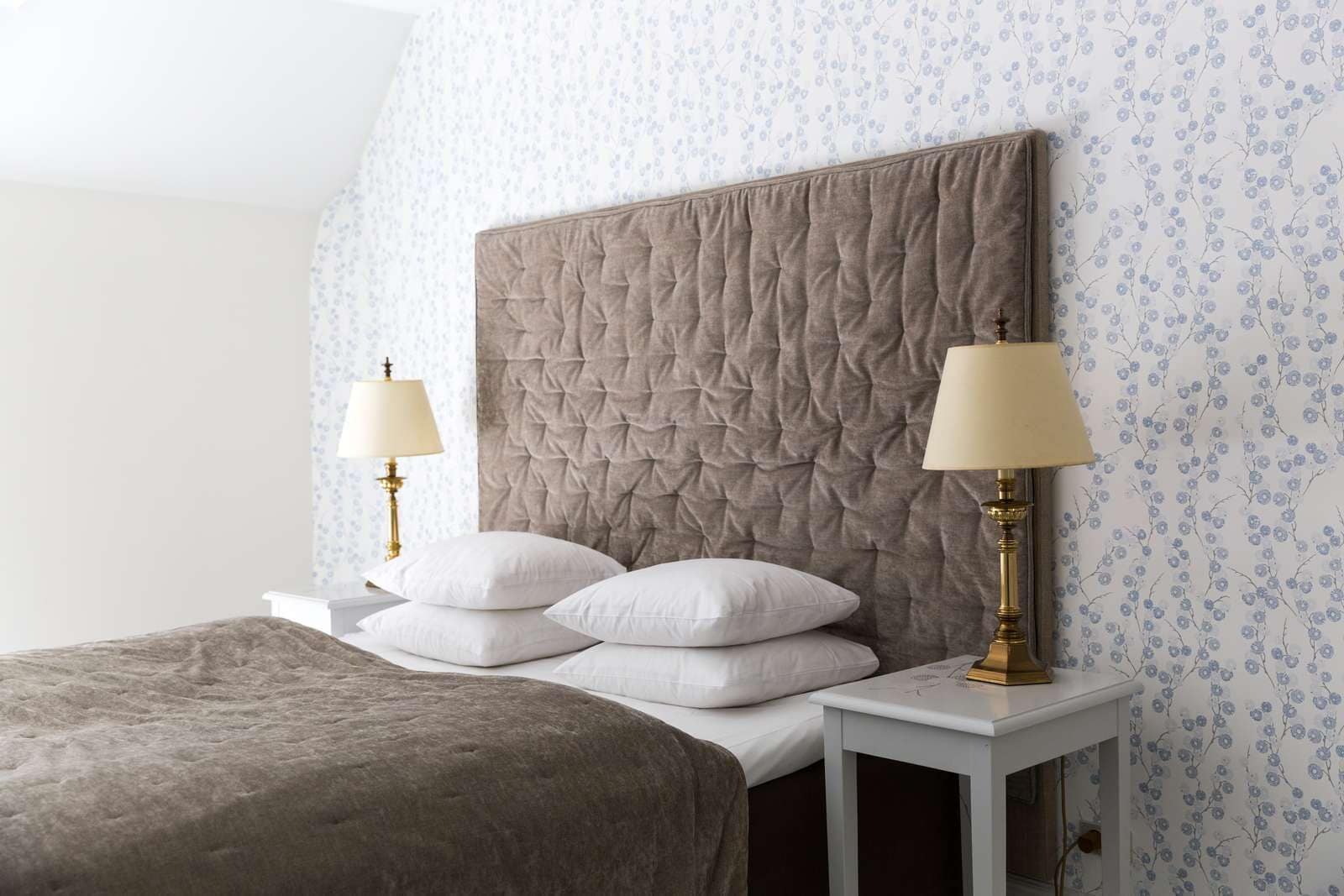 A neatly made bed with a large headboard, two bedside lamps, and a floral wallpaper pattern.
