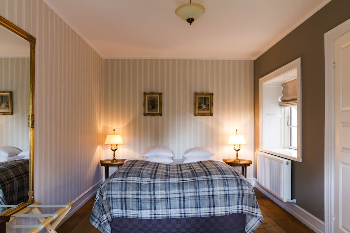 Une chambre d’hôtel avec un grand lit, deux tables de chevet avec des lampes, un miroir et une fenêtre donnant sur un beau paysage.
