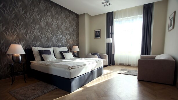 Ein modernes Schlafzimmer mit einem großen Bett, zwei Nachttischen mit Lampen, einem Sofa und Blick ins Freie durch ein Fenster mit Vorhängen.